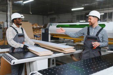 İki fabrika işçisi, ahşap panelleri tartışırken mobilya üretim tesisinde bir makinenin yanında dikiliyorlar.
