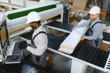 Bir mobilya fabrikasında mobilya üretimi için ahşap paneller kesen bir makineyle çalışan iki marangoz.