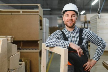 Marangoz güvenlik kaskı takıyor ve iş tulumu giyiyor. Bir mobilya fabrikasında ahşap çerçeveye gülümsüyor ve yaslanıyor.