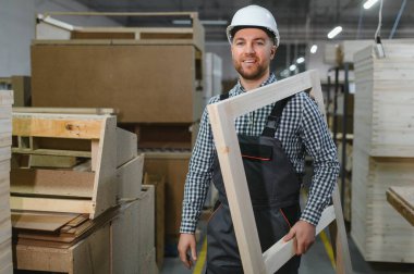 Gülümseyen marangoz mobilya fabrikasında ahşap çerçeve taşıyor, el işi ve üretim süreci sergiliyor.