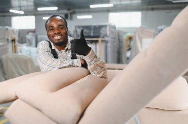 Afro-Amerikan işçiler mobilya fabrikasında kanepe monte ederken başparmak kaldırıyor. Arka planda tahta ve cnc makineleri görünür