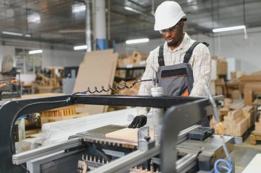 Marangoz güvenlik gözlüğü ve kask takıyor. Mobilya fabrikasında cnc makinası çalıştırıyor. Çalışma yüzeyine ahşap tahtalar yerleştiriyor.