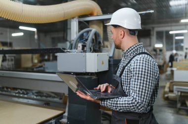 Marangoz koruyucu giysisi ve koruyucu giysisi var. Mobilya imalat fabrikasında dizüstü bilgisayarı olan bir Cnc makinesini programlıyor.