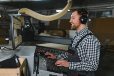 Marangoz kulak koruyucu takıyor. Mobilya fabrikasında cnc makinesi kullanıyor. Ahşap işleme programlıyor.