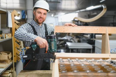 Marangoz güvenlik kaskı takıyor ve tulum giyiyor. Pnömatik çivi tabancası kullanıyor. Mobilya fabrikasında ahşap kanepe çerçevesi yapıyor.
