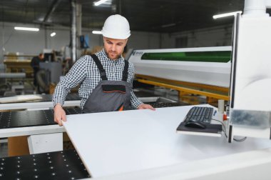 Marangoz güvenlik kaskı takıyor. Yoğun bir mobilya fabrikası ortamında modern bir cnc makinesine ahşap panel yerleştiriyor.