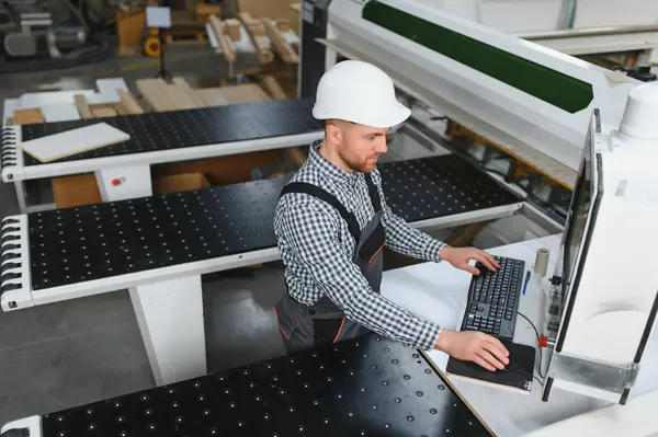 Mobilya fabrikasında güvenlik kaskı takan marangoz, işlemi bilgisayar aracılığıyla kontrol ediyor.