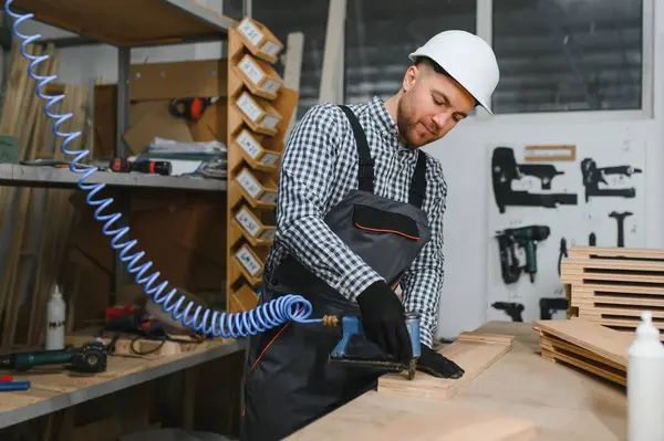 Marangoz güvenlik kaskı takıyor. Pnömatik çivi tabancası kullanıyor. Atölyede ahşap mobilya parçaları birleştiriyor.