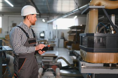 Bir mobilya fabrikasında güvenlik kaskı takan marangoz uzaktan kumandayla otomatik işlemi kontrol ediyor.