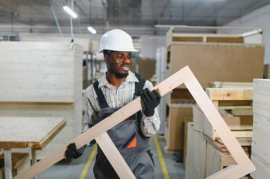 Gülümseyen Afro-Amerikan marangoz ahşap bir çerçeve tutuyor, işçilikle dolu bir mobilya fabrikasında özenle çalışıyor.