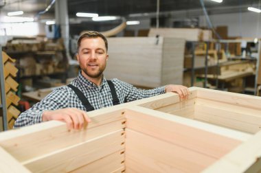 İşçi mobilya fabrikasında ahşap bir kanepe çerçevesi monte ediyor. Etrafı ahşap ve makinelerle çevrili, hassasiyet ve işçiliğe odaklanmış.