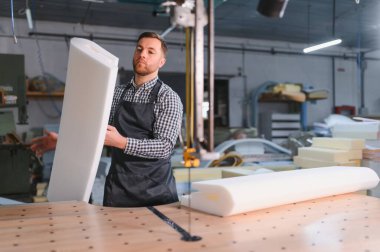 Önlük giymiş fabrika işçisi, bir mobilya üretim ortamındaki makine ve malzemelerle çevrili ahşap bir masada köpük parçaları tutuyor.