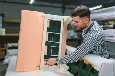 Marangoz bir mobilya fabrikasında ahşap bir kanepe çerçevesi monte ediyor, imalat sürecinde zanaat ve beceri sergiliyor.