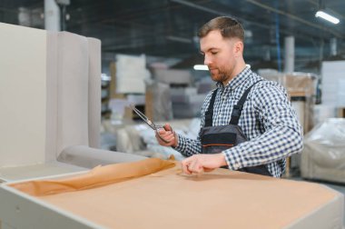 Marangoz mobilya fabrikasında çalışıyor. Kanepe monte ederken büyük bir makasla ambalaj kağıdı kesiyor.