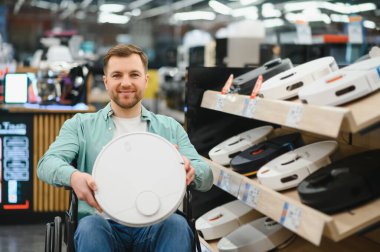 Tekerlekli sandalyede elinde elektrikli süpürgeyle gezen mutlu bir adam ev aletleri mağazasında alışveriş yapıyor.