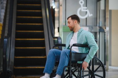 Tekerlekli sandalyede yürüyen merdivene bakan bir müşteri, arka planda erişilebilirlik işareti olan bir alışveriş merkezinde hareket sorunu yaşıyor.