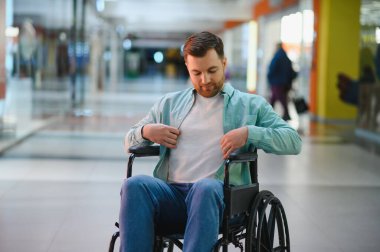 Tekerlekli sandalyede oturan sakallı adam, geniş bir alışveriş merkezinde gezinirken hafif gömleği ayarlıyor, perakende terapisinin keyfini çıkarıyor.