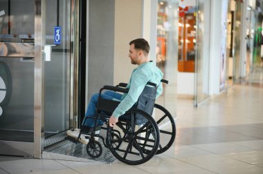 Engelli biri modern bir alışveriş merkezinde geziniyor, asansörden çıkarken tekerlekli sandalye rampası kullanıyor.
