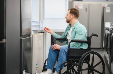 Tekerlekli sandalyedeki bir adam buzdolabının kapısını açıyor, yeni ev aletlerini seçiyor ve bir elektronik mağazasının çeşitli bağışlarında geziniyor.