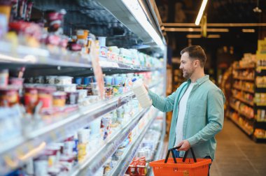 Süpermarkette mandıra ürünleri seçen bir adam, elinde bir alışveriş sepetiyle. Raflar koridor boyunca çeşitli yiyecek öğeleriyle dolu.