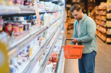 Modern bir süpermarketin soğutuculu bölümünden taze sebze seçerken elinde alışveriş sepeti tutan bir adam.