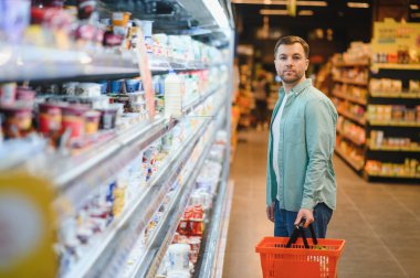 Elinde sepeti olan bir adam süpermarket koridorunda erzak seçiyor, mandıra ürünlerini ve temel ihtiyaçlarını aydınlatma altında keşfediyor.