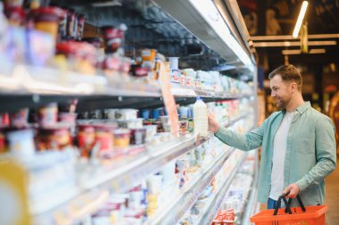 Süpermarketteki soğutuculu raftan bir şişe süt seçen gülümseyen adam elinde alışveriş sepeti tutuyor.