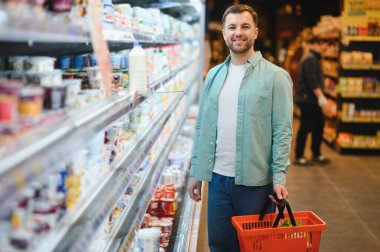 Cheerful man buying groceries in a supermarket, holding a shopping basket and smiling, walking along refrigerated shelves