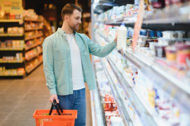 Müşteri marketin soğutuculu bölümünden yiyecek seçiyor, alışveriş sepeti tutuyor ve süte uzanıyor.
