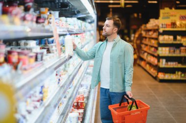 Süpermarketin soğutuculu bölümünden sebze seçen, alışveriş sepeti tutan ve bir şişe süte uzanan bir adam.