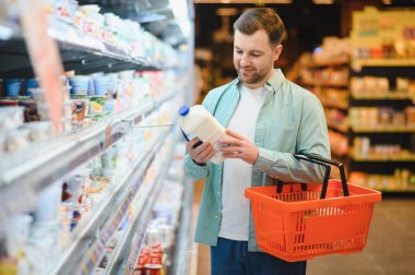 Elinde alışveriş sepeti olan ve markette süt kutusunu inceleyen bir adam. Raflar, arka planda çeşitli market eşyalarıyla dolu.
