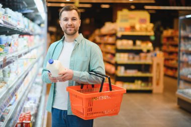 Smiling man holding milk bottle and shopping basket, buying groceries in supermarket dairy section