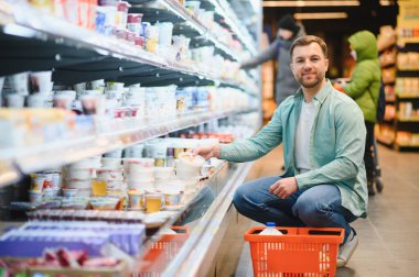 Man kneeling in a supermarket aisle, selecting dairy products from a shelf. He holds a basket filled with various groceries