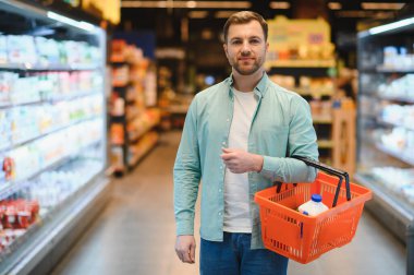 Man carrying a shopping basket filled with groceries, navigating the aisles of a busy supermarket while selecting fresh food items