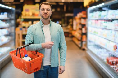 Man holding a shopping basket while selecting fresh groceries in a bustling supermarket, navigating the aisles filled with various products