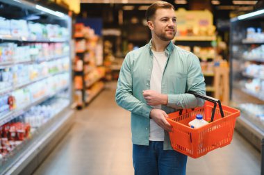 Bir marketin soğutuculu mandırasında yürürken alışveriş sepeti taşıyan bir adam taze ürünleri seçiyor.