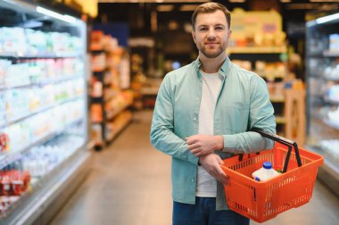 Man holding a shopping basket filled with groceries while navigating the aisles of a bustling supermarket, surrounded by various food options