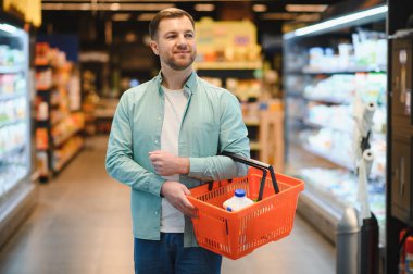 Man holding a shopping basket buying groceries in a supermarket, walking between shelves and choosing products