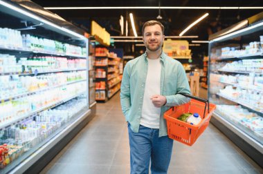 Smiling man doing grocery shopping holding a basket walking in supermarket between shelves full of refrigerated dairy products