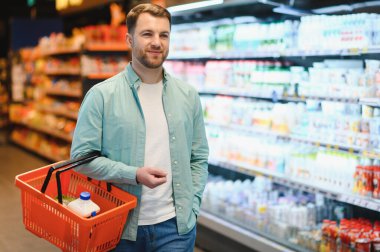 Gülümseyen bir adam alışveriş sepeti taşırken markette çeşitli ürünlerle dolu taze yiyecekler seçiyor.