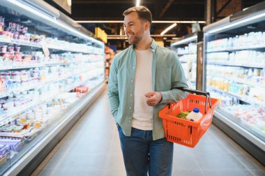 Süpermarket koridorlarında yürürken alışveriş sepeti taşıyan mutlu bir adam taze ürünler seçiyor ve sağlıklı bir yaşam tarzının tadını çıkarıyor.