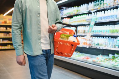 Süpermarkette mandıra ürünleri reyonunda yürürken elinde sebzelerle turuncu alışveriş sepsiyonu taşıyan bir adam.