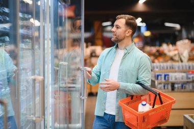 Dondurulmuş bir koridordan sebze ve süt dolu bir alışveriş sepetini parlak ışıklı bir süpermarketten seçen bir adam.
