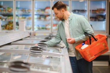 Adam markette dondurulmuş ürünler seçiyor, elinde süt ve sebzelerle alışveriş sepeti tutuyor.