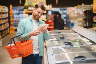 Alışveriş sepeti tutan ve marketteki dondurulmuş ürünlerin malzemelerini kontrol eden adam.