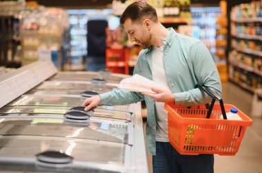 Süpermarketten yiyecek seçen bir adam, elinde yiyecek dolu bir sepet, buzdolabı bölümünden taze ürünler seçmeye odaklanmış.