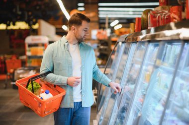 Bir adam dondurulmuş ürünleri marketten seçiyor, taze ürünlerle dolu bir alışveriş sepetini tutuyor ve soğutucu koridorlarda geziniyor.