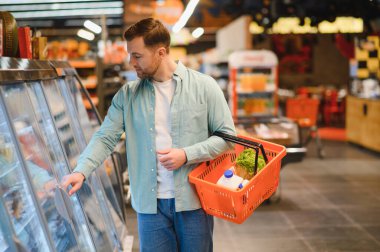 Süpermarketin soğutulmuş bölümünden yiyecek seçen bir adam taze ürünlerle dolu bir alışveriş sepeti tutuyor.