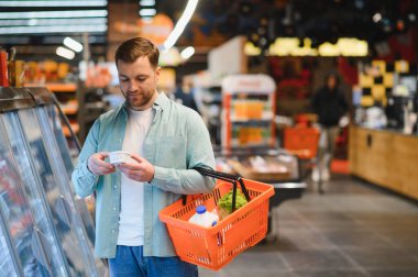 Alışveriş sepeti tutan ve marketin soğutulmuş koridorundaki yoğurt malzemelerini kontrol eden adam.