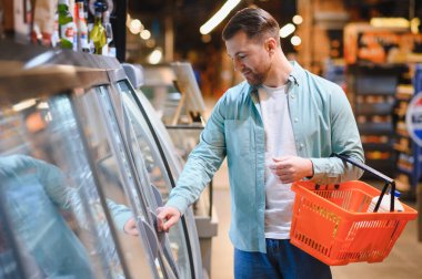 Süpermarketin soğutulmuş bölümünden yiyecek seçen bir adam, elinde alışveriş sepeti tutarken taze ürünleri ve süt ürünlerini dikkatle inceliyor.